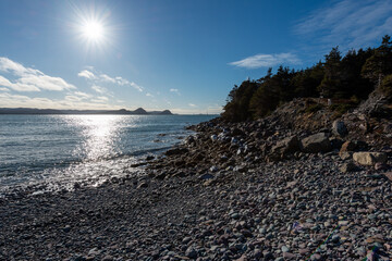 The smooth crystal clear ocean water under a blue sky with clouds. The shoreline is covered in...