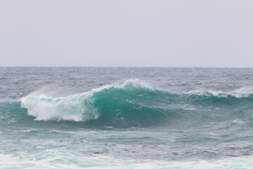 An angry turquoise green color massive rip curl of a wave as it barrels rolls along the ocean. The white mist and froth from the wave are foamy and fluffy. The ocean in the background is deep blue.