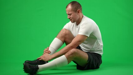 Troubled Caucasian sportsman sitting on green screen holding injured leg. Side view wide shot of sad young handsome man with painful sport trauma at chromakey background