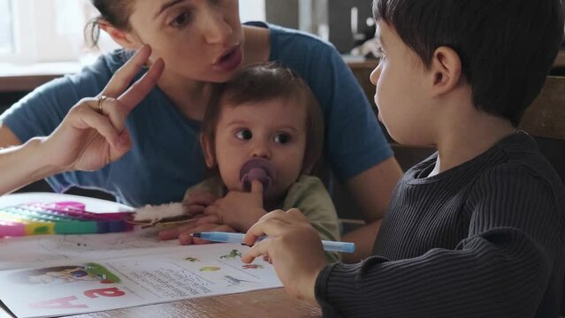 Little boy doing his math homework with a little help of his mother. The mother explains the math problem to her son , while she is also worried about her baby