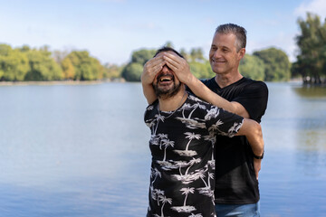 Naklejka premium Couple of mature gay men, one surprising the other by covering his eyes in a lake. Concept of surprise, happiness, game
