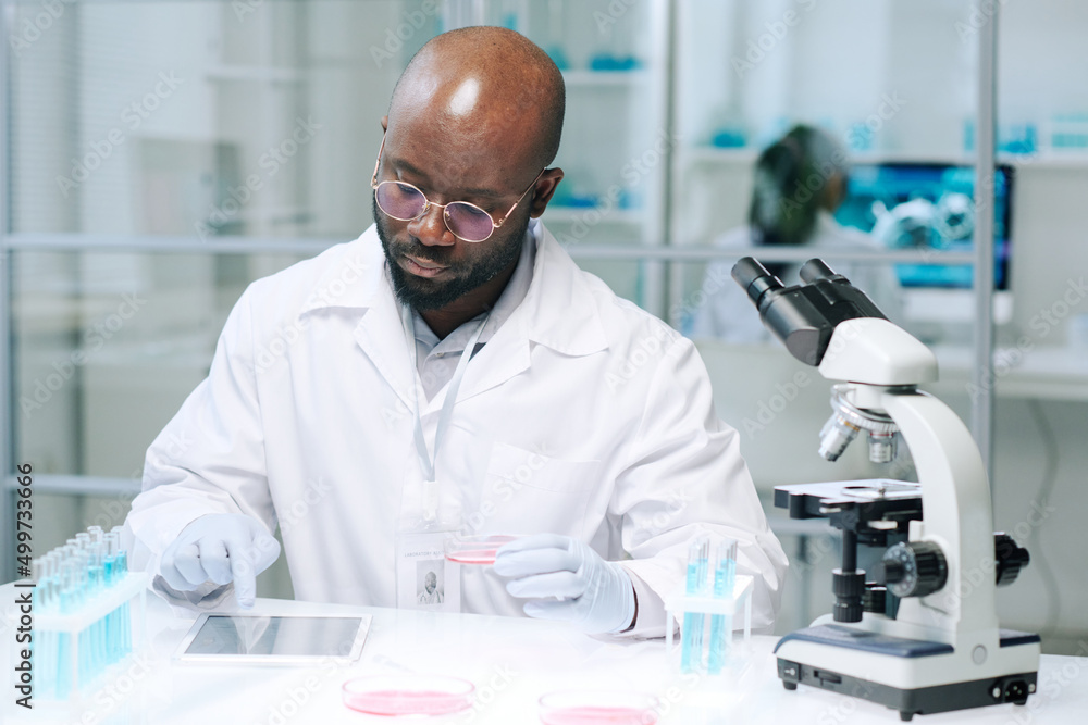 Poster african american male scientist sitting by workplace in laboratory and scrolling in tablet while sea