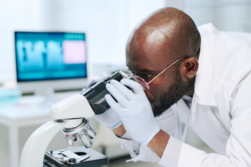 Young serious black man in whitecoat and gloves working with microscope while making scientific...