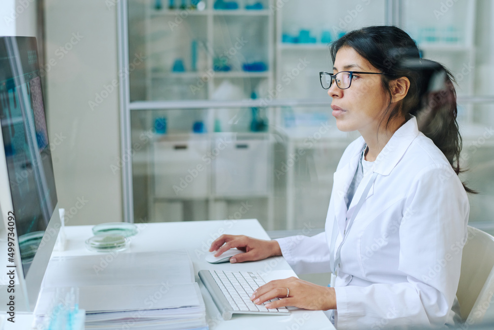 Aufkleber young pretty female scientist in whitecoat and eyeglasses using computer for studying new virus whil