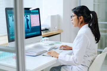 Obraz na płótnie Canvas Side view of young Hispanic female researcher sitting in front of computer monitor and studying new virus in modern scientific laboratory