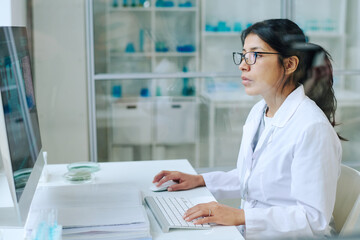 Young pretty female scientist in whitecoat and eyeglasses using computer for studying new virus...