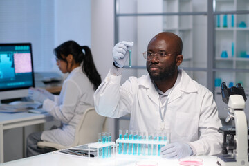 African American male virologist in gloves and lab coat looking at blue liquid in flask against...