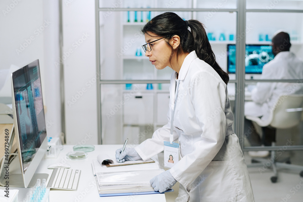 Aufkleber Side view of young serious female clinician looking at computer screen with coronavirus macro image while making notes in document