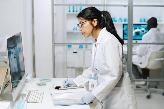 Side view of young serious female clinician looking at computer screen with coronavirus macro image while making notes in document