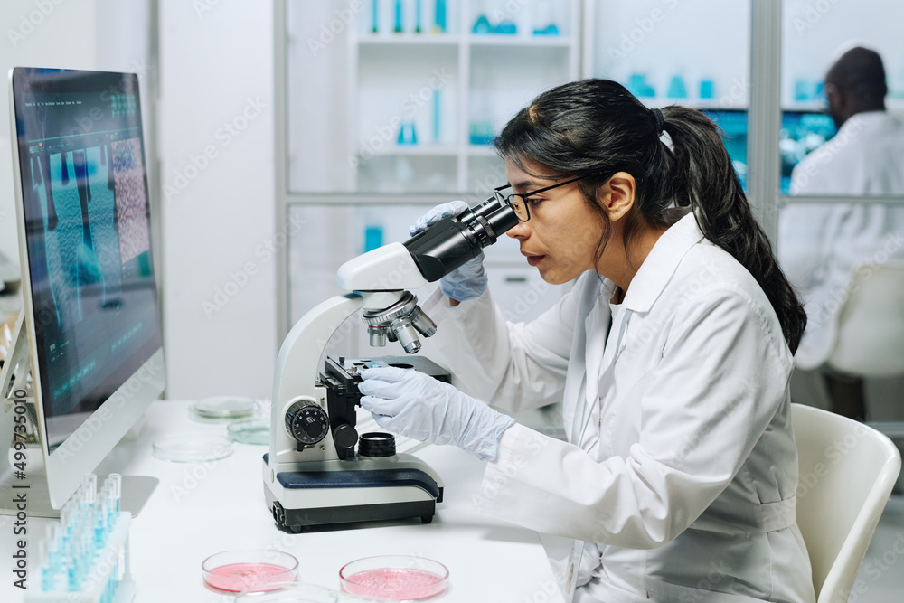 Poster side view of female biochemist in whitecoat, gloves and eyeglasses looking in microscope while study
