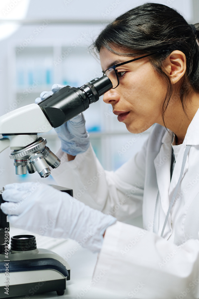 Poster young serious female microbiologist or virologist using microscope in laboratory while making scient