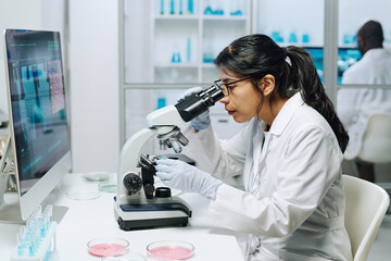 Side view of female biochemist in whitecoat, gloves and eyeglasses looking in microscope while...