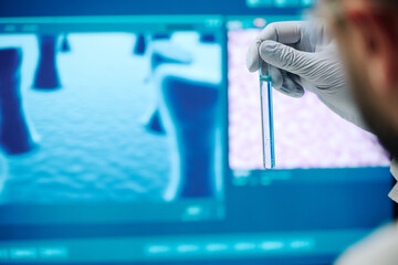 Flask with blue liquid in gloved hand of contemporary male virologist or researcher holding it against computer screen with covid molecule