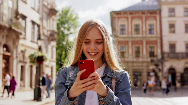 Young blonde woman tapping and scrolling on mobile phone outdoors. Beautiful girl texting message on smartphone and chatting at street. Female uses a smartphone. View photos and videos on the phone.