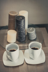 Two white cups of hot aromatic coffee on a brown table with candles. Composition in brown and beige tones.