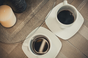 Two white cups of hot aromatic coffee on a brown table with candles. Composition in brown and beige...