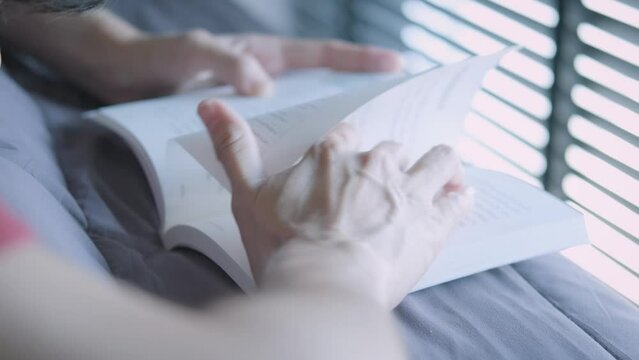 Hands of short-haired Asian woman opening textbooks Resting on a bed by the window with wooden blinds that have enough light during the day to protect the eyes. Concept of studying at home covid-19