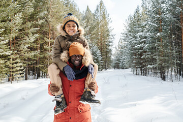 Fototapeta na wymiar Happy young black man in winterwear carrying his cute son on shoulders while spending time in th forest among evergreen trees