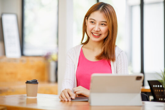 Portrait of a beautiful young and intelligent-looking Japanese Asian woman student wearing a white shirt and pink tracker smiling as she works on her laptop in a university classroom.