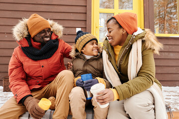 Cute happy boy in winterwear sitting between his parents on porch of their family country house, having hot tea and chatting