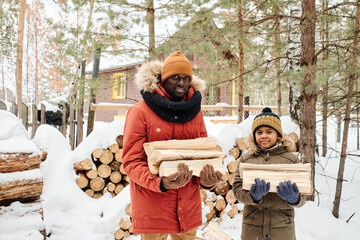 Happy young African American father and son in warm winter jackets and beanies carrying firewood to their country house for heating it