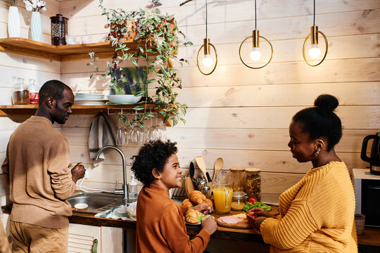 Cute little boy helping him mother with cooking breakfast and talking to her while both standing by kitchen table inside country house