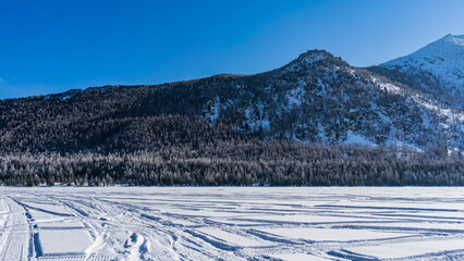 Tire tracks are visible on the frozen and snow-covered lake. The coniferous forest on the shore is covered with frost. A mountain range against the blue sky. Altai. Lower Multinskoe Lake