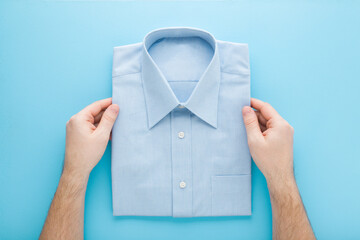 Young adult man hands holding folded classic shirt on light blue table background. Pastel color....