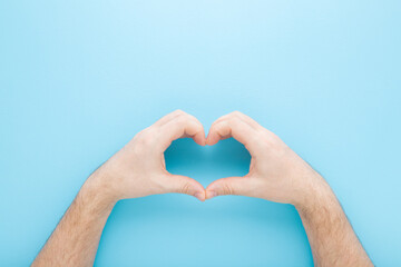 Heart shape created from young adult man hands on light blue table background. Pastel color. Love and happiness concept. Closeup. Point of view shot. Top down view.