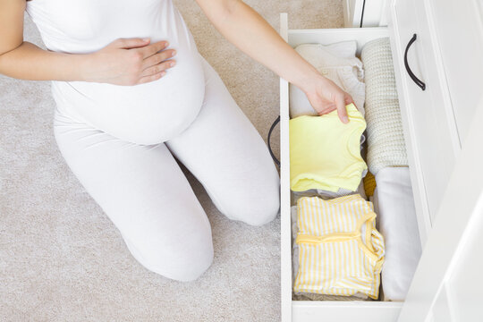Young adult pregnant woman with big belly sitting on carpet at nursery room. Hand holding bodysuit and sorting clothes for future baby in white drawer box. Preparing things in pregnancy time.