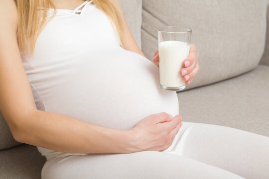 Young adult pregnant woman in white clothes sitting on sofa and holding glass of fresh milk at home. Healthy drink in pregnancy time. Baby expectation. Side view.