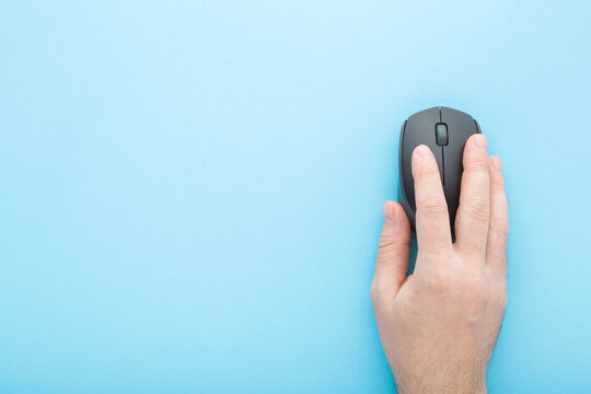 Young adult man hand using dark black computer optical mouse on light blue table background. Pastel color. Closeup. Empty place for text. Top down view.