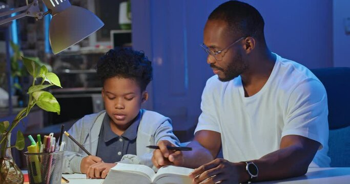 African American Businessman Father Teaching And Helping Son Doing Homework In Living Room At Late Night. Home Studying At Distance Learning, Family Spending Quality Time Together. Lifestyle Concept.