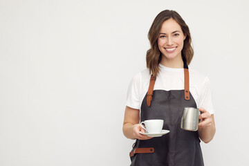 Beautiful young barista woman looking in camera and smiling, while serving a cup of coffee and holding a milk jug. Isolated on white background.	 - obrazy, fototapety, plakaty