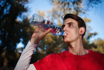 Young sportsman drinking water from bottle after running and listening songs while jogging. Body hydration.