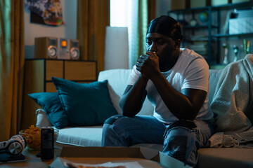 Fototapeta premium Sad hurt young man sits in living room at night, guy can't sleep contemplates, room lit by street light coming through window. The boy is heartbroken, eating nerves with pizza and chips.