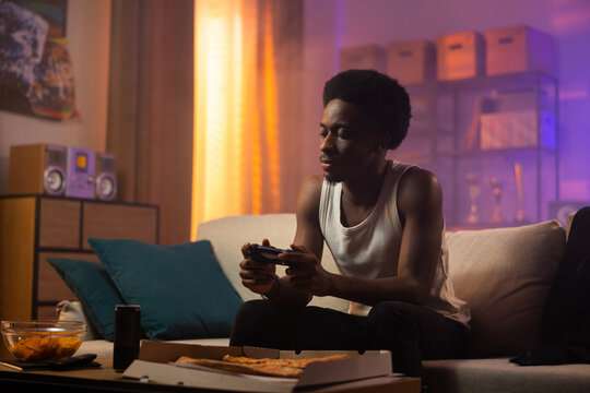 An African American man relaxes on the living room couch playing video games using a gamepad, a young gamer relaxes at home engaged in digital virtual activity