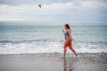 A plump woman in a bathing suit enters the water during the surf. Alone on the beach, Gray sky in the clouds, swimming in winter.