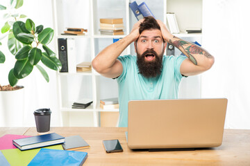 Surprised man with jaw dropped clutching head sitting at laptop in office, surprise