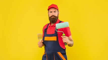 bearded man painter in work clothes hold paint roller and brush on yellow background