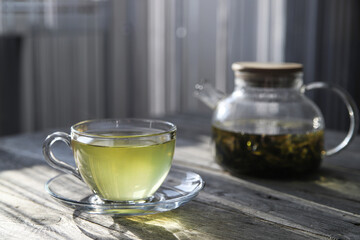 Green tea in a glass cup and teapot on a wooden table in the kitchen on a sunny morning. Hot healthy drink. Atmospheric breakfast background