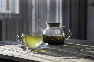 Green tea in a glass cup and teapot on a wooden table in the kitchen on a sunny morning. Hot healthy drink. Atmospheric breakfast background