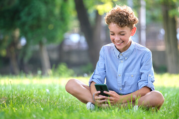Naklejka na ściany i meble Young boy browsing his smartphone outdoors in summer park. Using cellphone for leasure and education concept