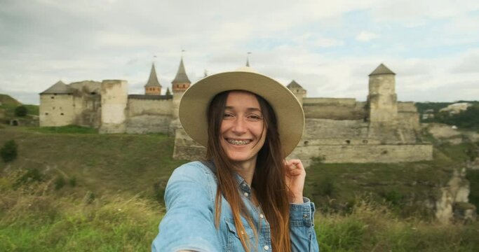 young woman in a hat looks at the camera in front of an ancient castle with towers. Daytime, cloudy, medium shot. Take a selfie in front of you