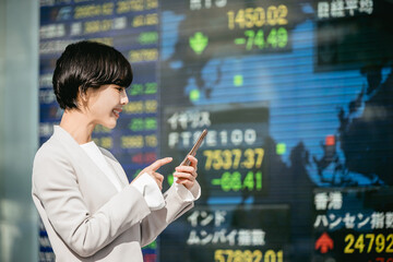 株価ボードの前でスマートフォンを見る女性