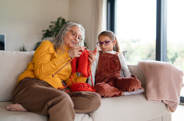 Little girl sitting on sofa with her grandmother and learning to knit indoors at home.