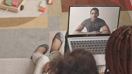 From above slowmo of young Black military man communicating with his beloved family through video chat on laptop, talking to little daughter and wife sitting on sofa at their home
