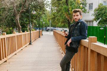 Fototapeta premium Posing, the young bearded handsome man is wearing a denim jacket, She leans her back on the bridge and poses for the camera, has a serious facial expression