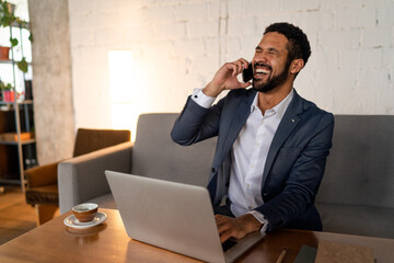 Cheerful young businessman sitting on sofa and talking on cellphone in office.