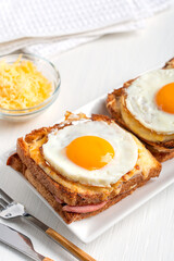 Homemade Croque Madame French hot sandwiches made of ham and cheese inside toasted bread topped with fried egg with yolk served for breakfast on plate with tableware and towel on white wooden table 